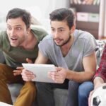 Três amigos assistem a um jogo de futebol com expressões de surpresa e animação, um deles apontando para algo fora da tela, outro segurando um tablet, e o terceiro segurando uma bola de futebol e todos descobrindo como investir em clubes de futebol.