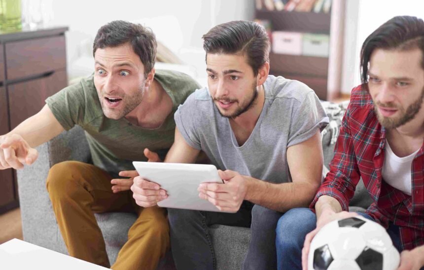 Três amigos assistem a um jogo de futebol com expressões de surpresa e animação, um deles apontando para algo fora da tela, outro segurando um tablet, e o terceiro segurando uma bola de futebol e todos descobrindo como investir em clubes de futebol.