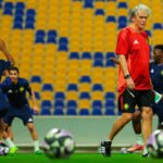 Jorge Jesus, técnico de futebol de cabelos brancos e camisa vermelha, caminha em campo gramado durante um treino do Al-Nassr, com jogadores em uniformes azuis ao fundo e bolas de futebol em primeiro plano.