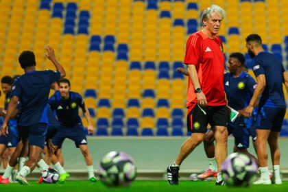 Jorge Jesus, técnico de futebol de cabelos brancos e camisa vermelha, caminha em campo gramado durante um treino do Al-Nassr, com jogadores em uniformes azuis ao fundo e bolas de futebol em primeiro plano.