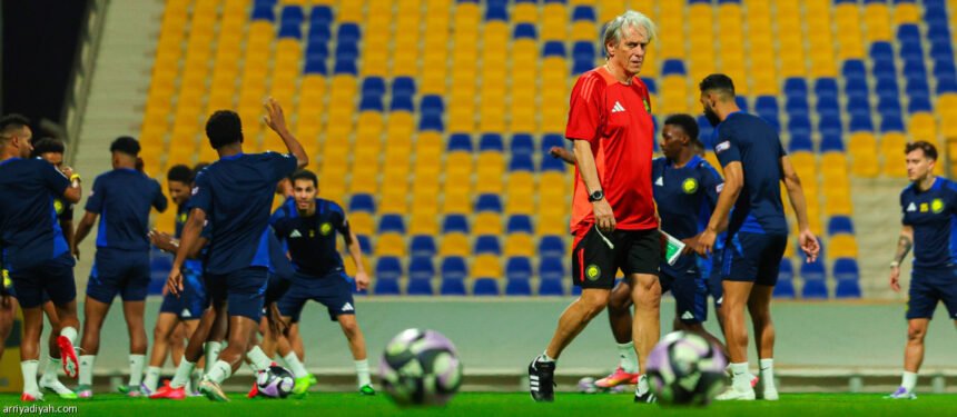 Jorge Jesus, técnico de futebol de cabelos brancos e camisa vermelha, caminha em campo gramado durante um treino do Al-Nassr, com jogadores em uniformes azuis ao fundo e bolas de futebol em primeiro plano.